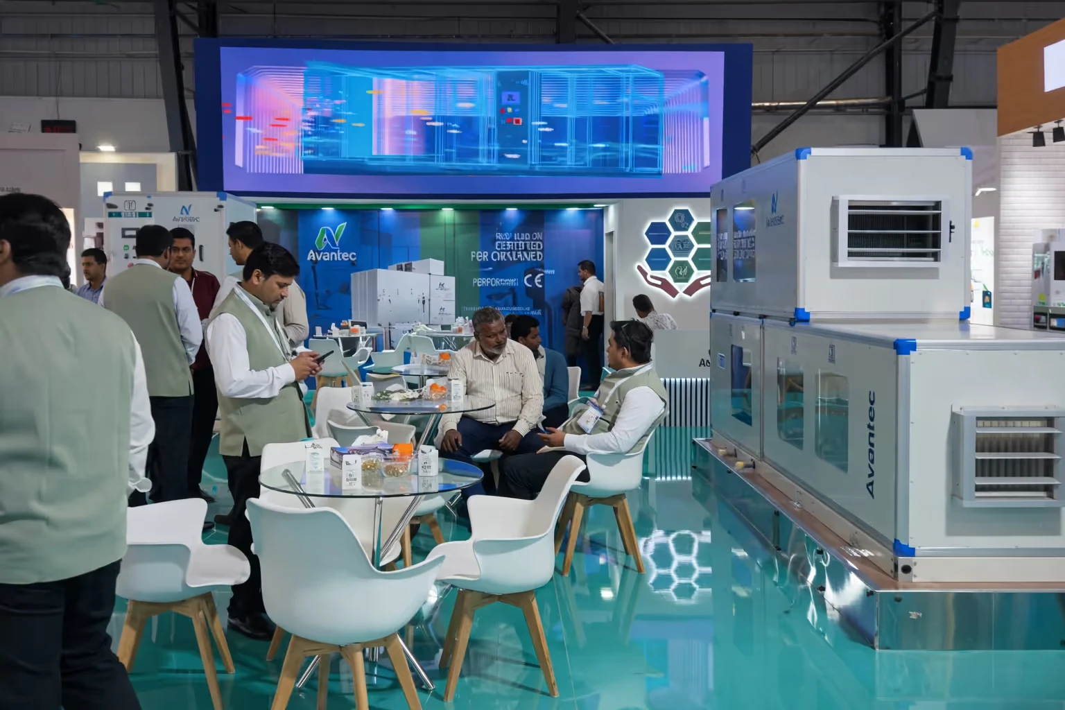Trade show booth with people seated and standing, featuring Avantec branded industrial equipment and a digital display of a cooling system.