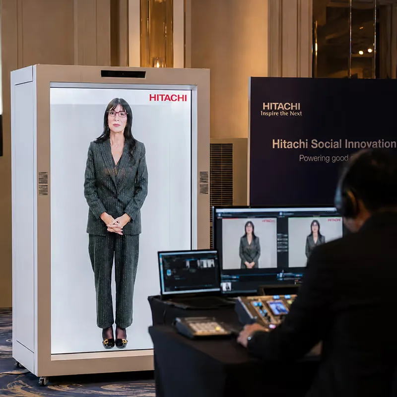 HoloBox911 unit on stage in front of a Hitachi Social Innovation Powering good backdrop, with a crew member alongside, at Hitachi's company event