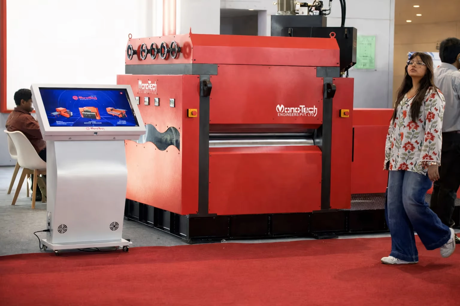 Large red industrial machine with a control panel touchscreen display on the left and a woman walking nearby in a floral shirt and wide jeans.