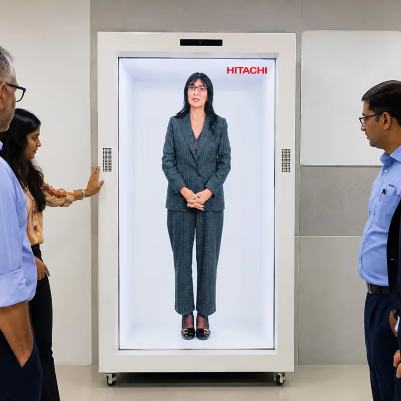 Delegates walking up to and interacting with the HoloBox911 holographic display unit in a lit lobby area at the Hitachi corporate event in New Delhi