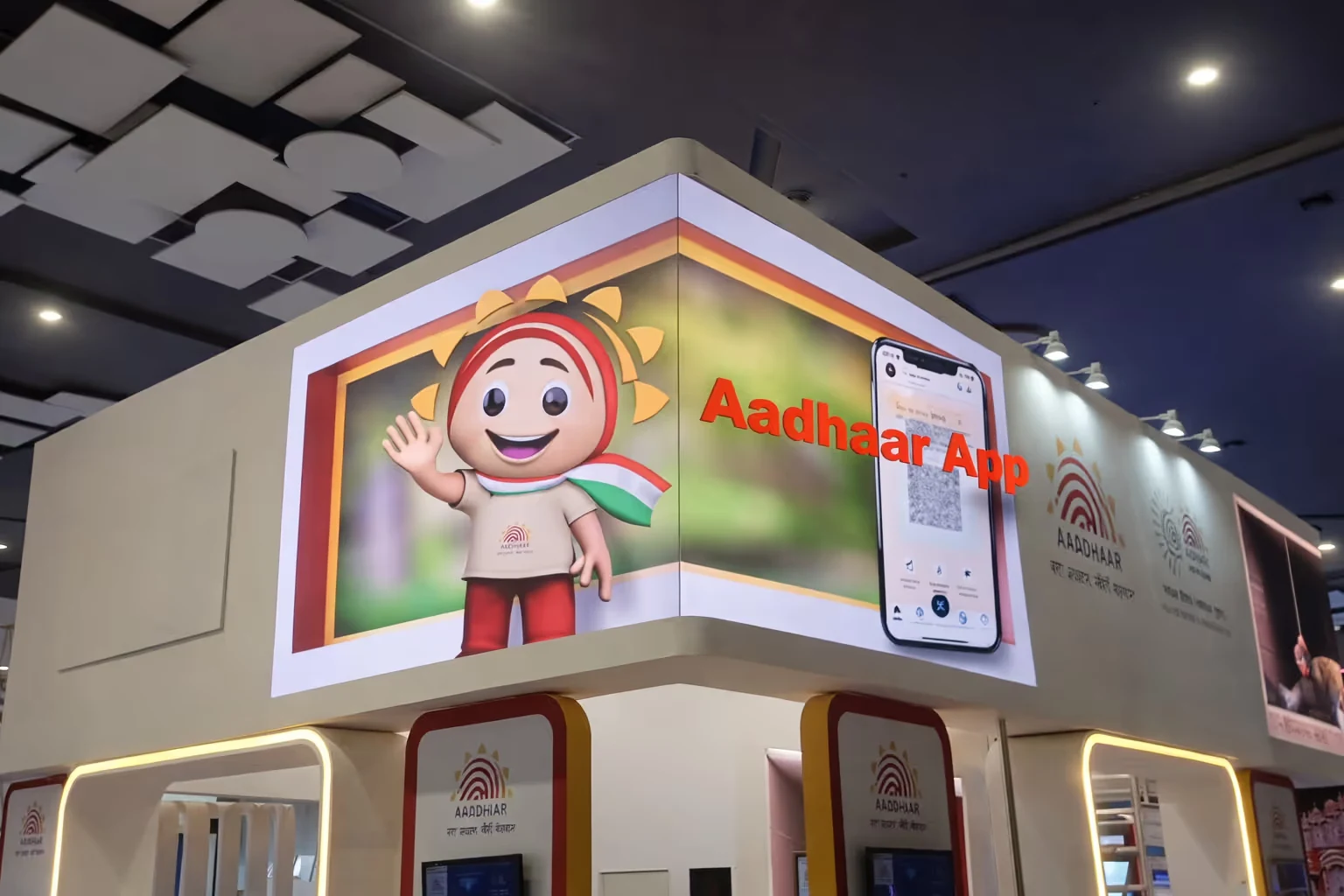 Exhibition booth featuring a cartoon sun mascot with Indian flag scarf and a digital display promoting the Aadhaar App.