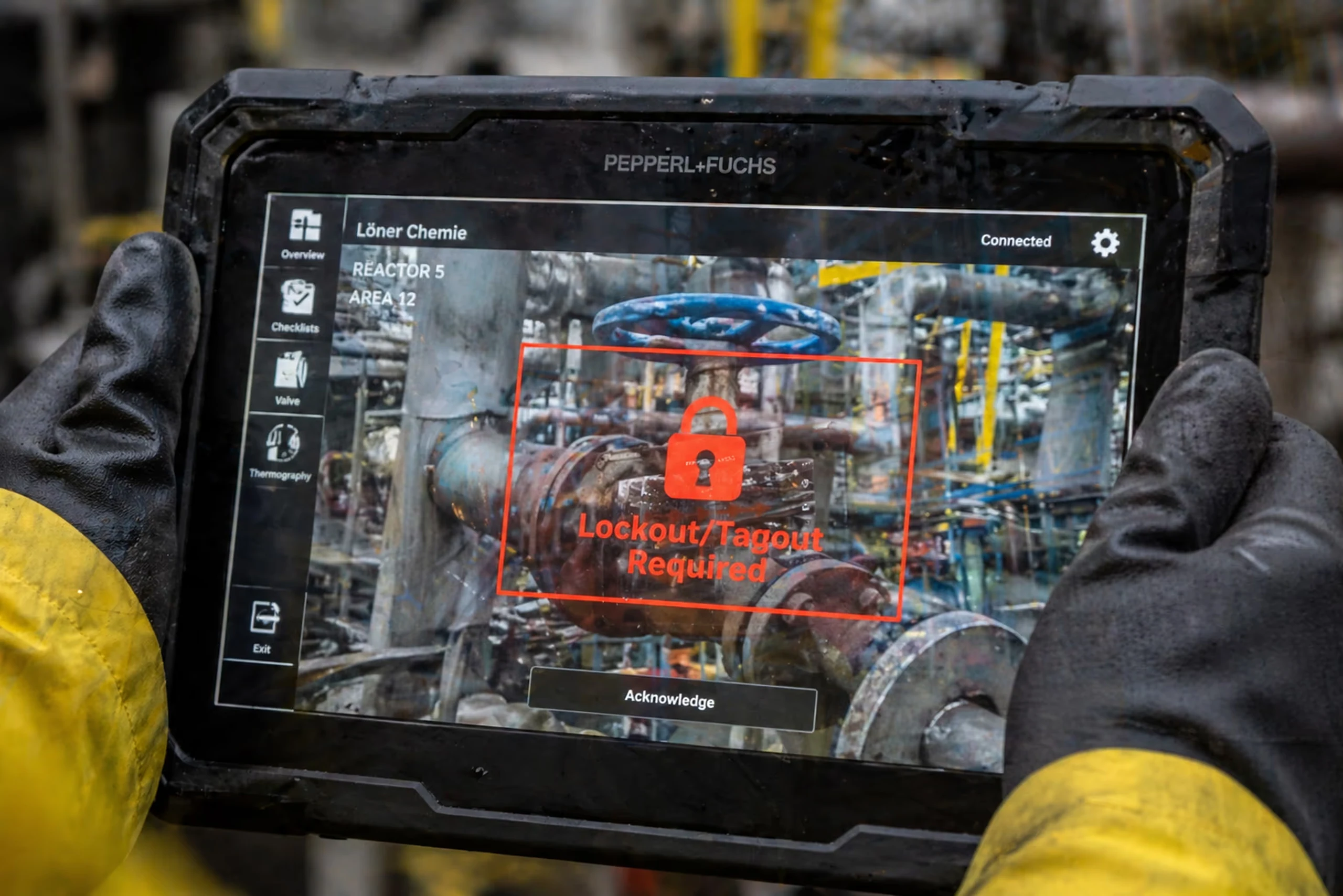 Industrial worker wearing gloves holding a rugged tablet displaying a red lock warning with 'Lockout/Tagout Required' over a valve in a chemical