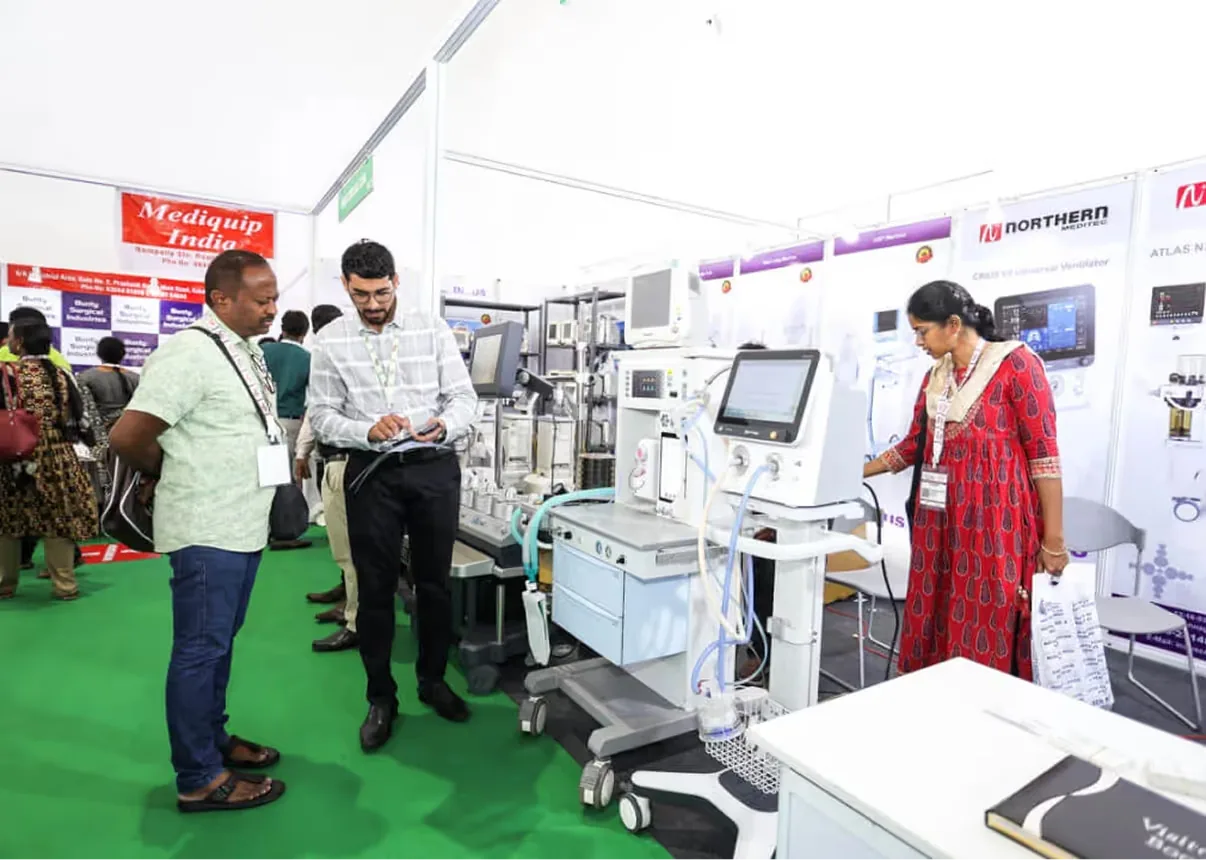Exhibition booth photo of visitors examining a medical ventilator and diagnostic equipment at the Mediqip India stand, with a sales