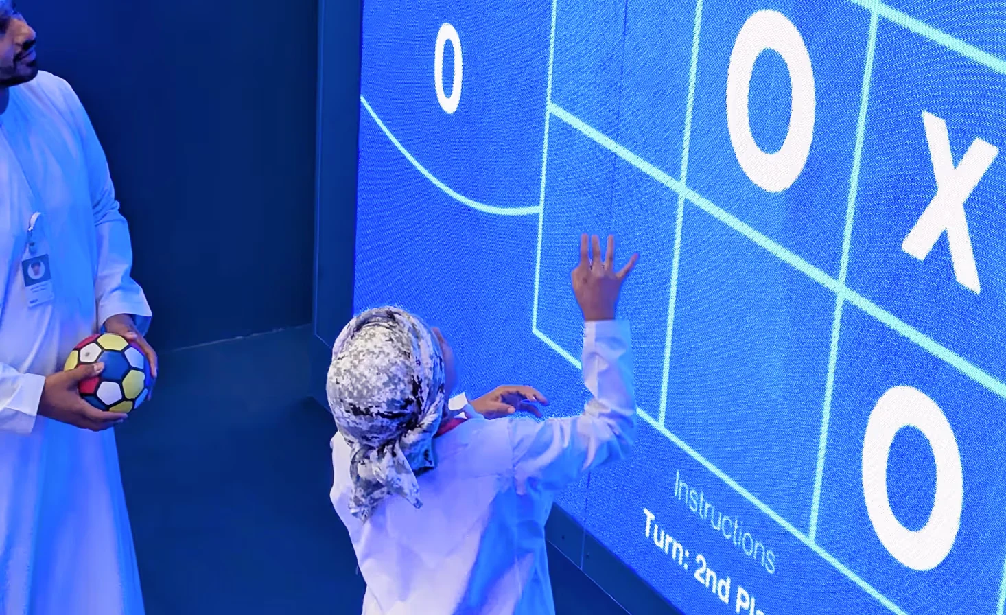 Visitor in traditional Omani attire throwing a ball at an XWall interactive tic-tac-toe game, with a second player waiting to take their turn
